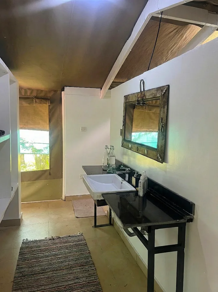 A rustic yet modern bathroom inside a canvas safari tent at Kaboso Safari Camp, featuring a black vanity with a white vessel sink, a timber-framed mirror, and a small woven rug on the floor. Sunlight streams in through a mesh window section, illuminating the white walls and sloped tent ceiling.