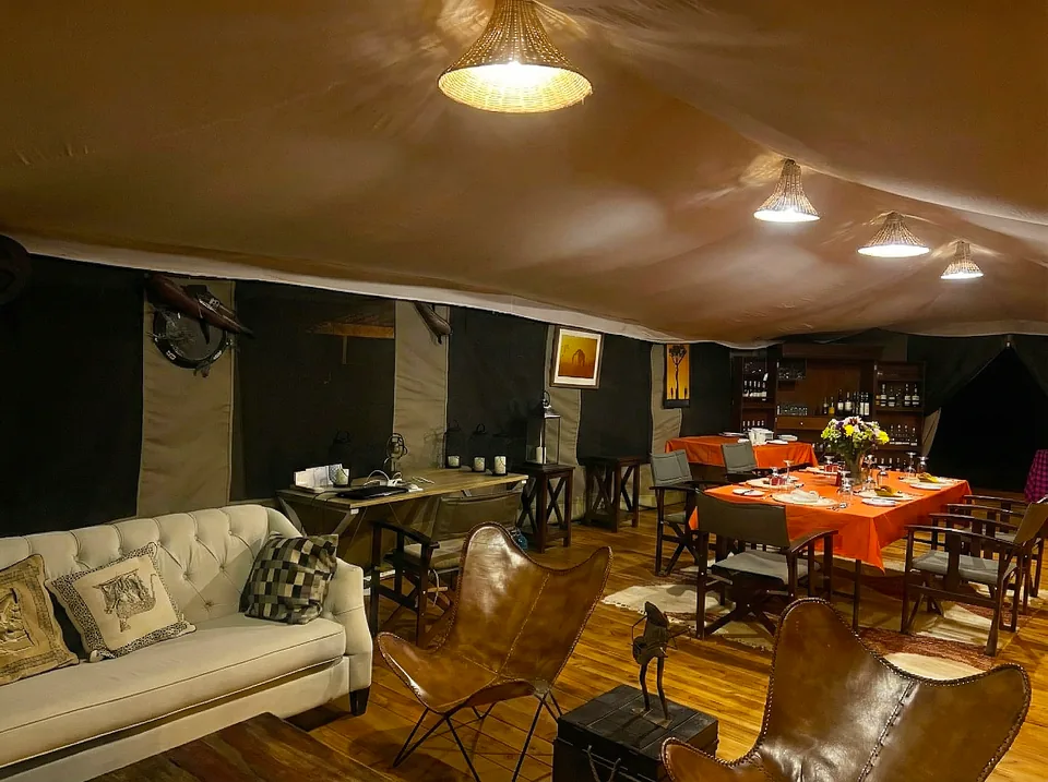 A spacious safari tent interior at Kaboso Safari Camp serving as a combined lounge and dining area, warmly lit by woven pendant lamps. It includes a beige tufted sofa with patterned throw pillows, brown leather butterfly chairs, and dining tables set with vibrant orange tablecloths and tableware.