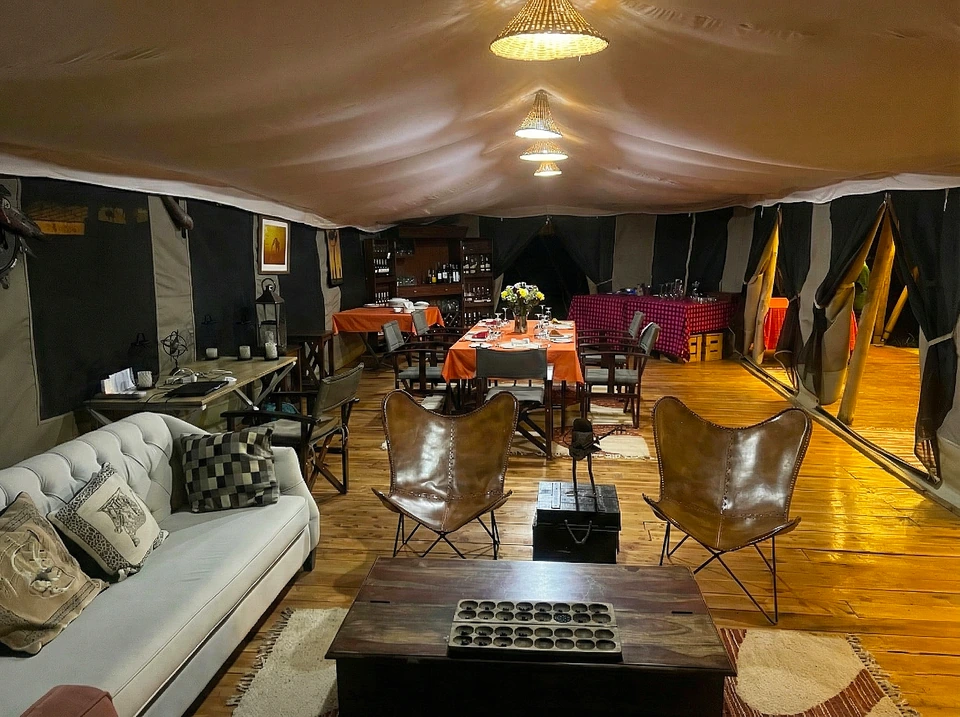 An expansive interior view of a luxury safari dining and lounge tent at Kaboso Safari Camp. It features a light grey tufted sofa, brown leather butterfly chairs, dining tables with red-checked and orange tablecloths, and a traditional Mancala-style game board placed on a wooden coffee table in the foreground.