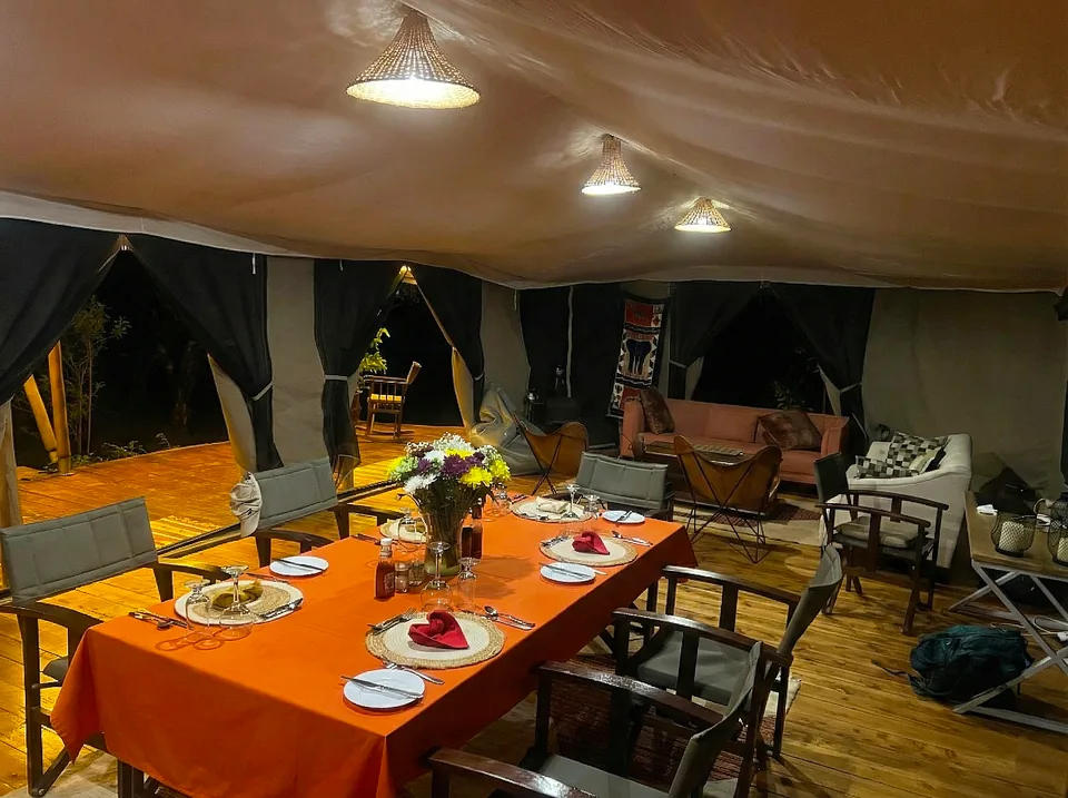 An elongated dining table set for dinner under a canvas tent at Kaboso Safari Camp at night, layered with a bright orange tablecloth and a central floral arrangement. The space is warmly illuminated by multiple woven pendant lamps, with various chairs including modern grey armchairs gathered around the table.