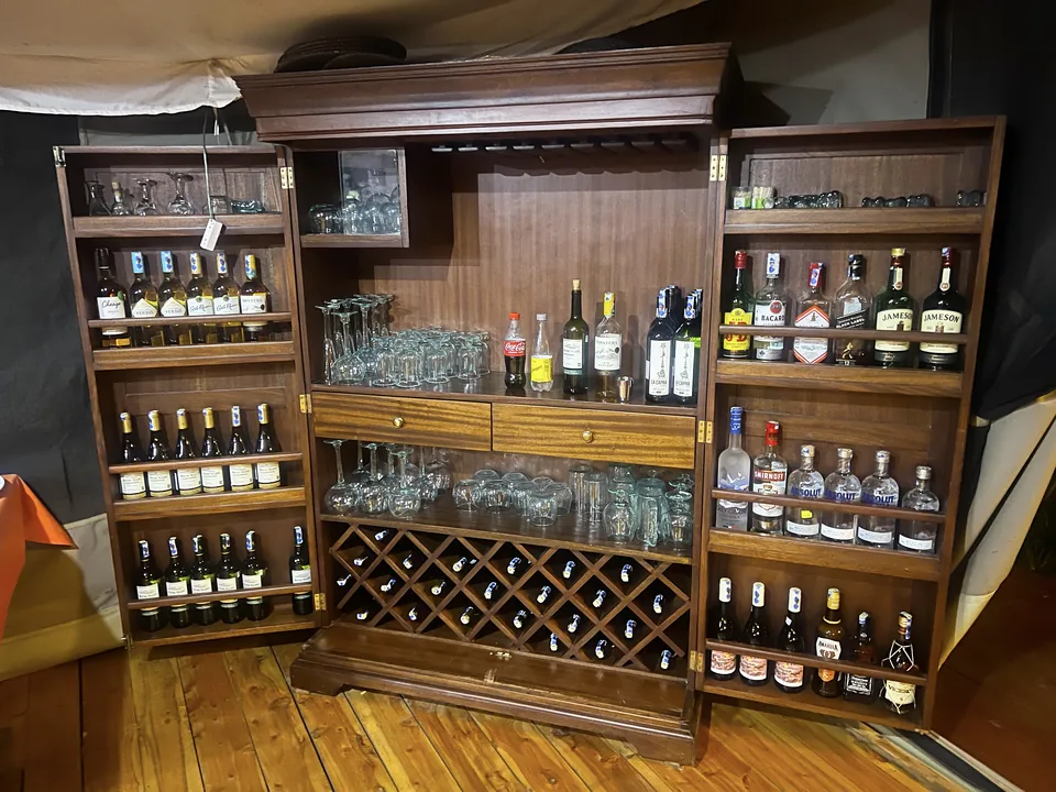 A large, open wooden bar cabinet inside a tent setting at Kaboso Safari Camp, displaying a wide assortment of wine and liquor bottles on multiple shelves. The furniture piece includes upper open shelving for glassware, two central drawers, and a built-in cross-hatch wine rack storing bottles horizontally at the bottom.