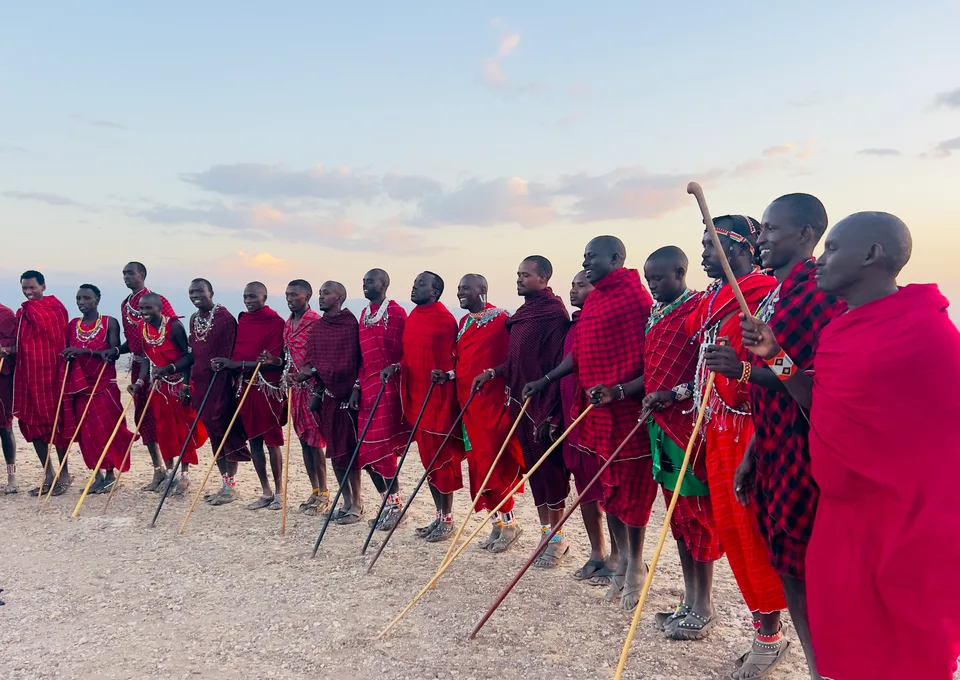 Group of Maasai men in red shukas standing in a line during a cultural performance at sunset.